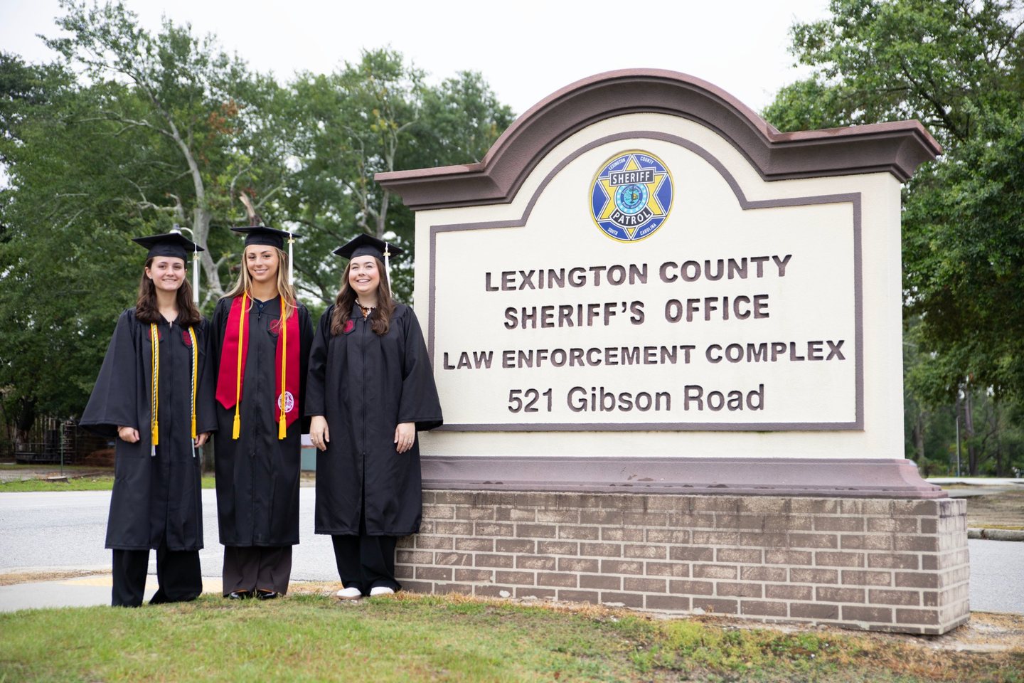Three USC graduates in caps and gowns beside the Lexington County Sheriff&rsquo;s Office sign