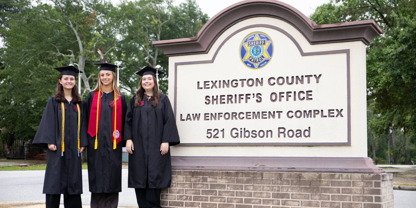 Three University of South Carolina graduates in caps and gowns beside the Lexington County Sheriff’s Office sign